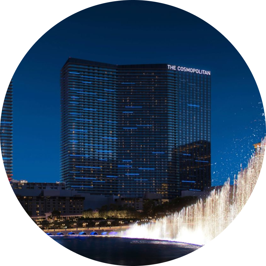 The image features The Cosmopolitan hotel, clearly identified by illuminated signage on the building. The scene is set at night, with the hotel’s modern glass facade reflecting city lights. In the foreground, a dramatic water fountain display is visible, adding dynamic movement and visual interest to the urban landscape.